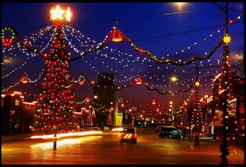 A festive street scene adorned with colorful Christmas lights and decorations, glowing under a twilight sky.