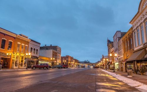 A quiet downtown street at dusk, lined with historic buildings and shops, reflecting lights on wet pavement.