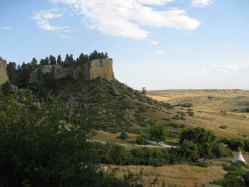 A scenic landscape featuring a rocky cliff, green hills, and a clear blue sky with scattered clouds.