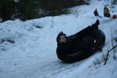 A person joyfully sledding down a snowy hill on a black inner tube, surrounded by snow and trees.