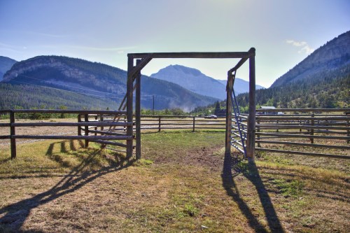 A wooden gate opens to a scenic view of mountains and a grassy field under a clear blue sky.