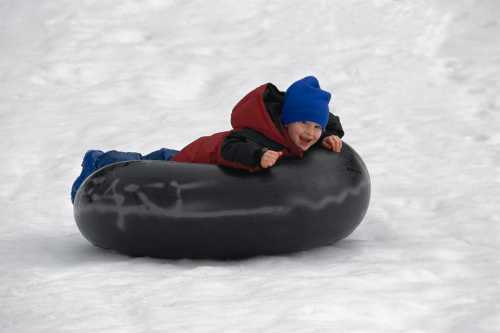 A joyful child in a blue hat and red jacket lies on a black snow tube, sliding down a snowy slope.