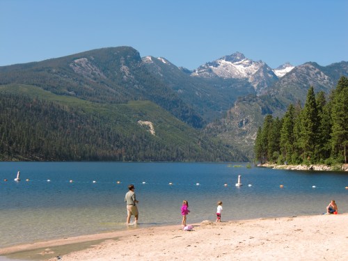 A sandy beach by a clear lake, with mountains in the background and people enjoying the water on a sunny day.