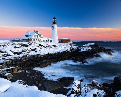 A snowy lighthouse stands on rocky shores, with waves crashing and a colorful sunset in the background.