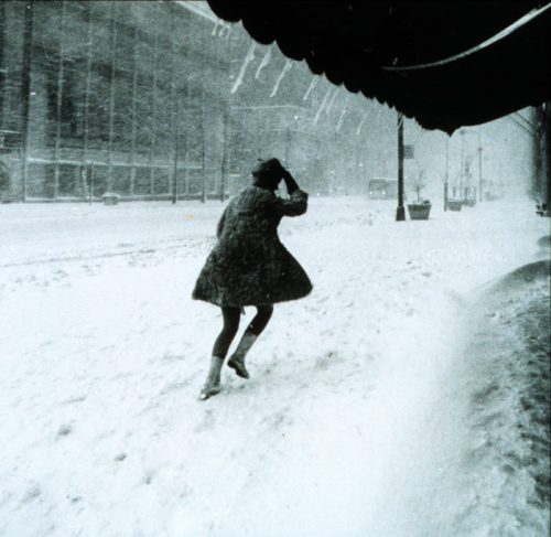 A person struggles against a heavy snowstorm, with snow covering the street and icicles hanging from a nearby awning.