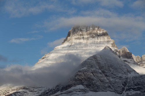 A snow-capped mountain peak rises against a blue sky, partially shrouded in clouds.