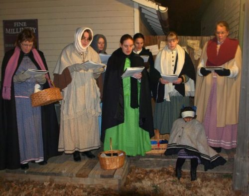 A group of women in historical clothing singing outdoors, with baskets and a child in a shawl nearby.
