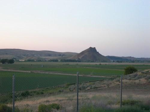 A distant view of a rocky hill surrounded by green fields under a pastel sky, with a fence in the foreground.
