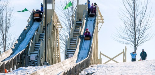 Two snow slides with people riding down, surrounded by trees and a clear blue sky. Spectators watch from the side.