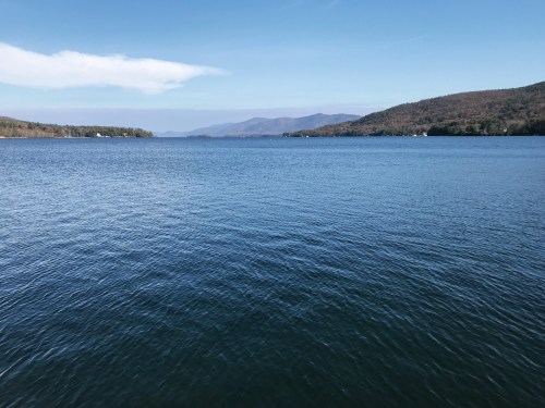 A serene lake view with calm blue water, surrounded by mountains and a clear blue sky.