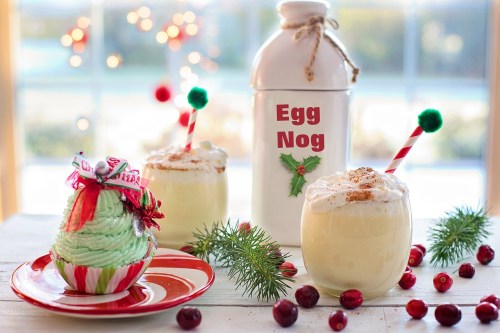 Festive scene with eggnog in glasses, a cupcake, and a decorative milk bottle, surrounded by holiday decorations.