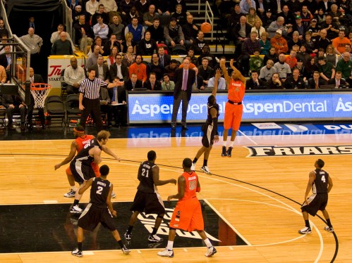 A basketball player in an orange jersey shoots a ball while teammates and opponents watch on a crowded court.