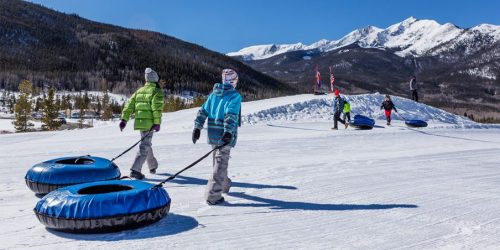 People walking on snow with inflatable tubes, surrounded by mountains and a clear blue sky.