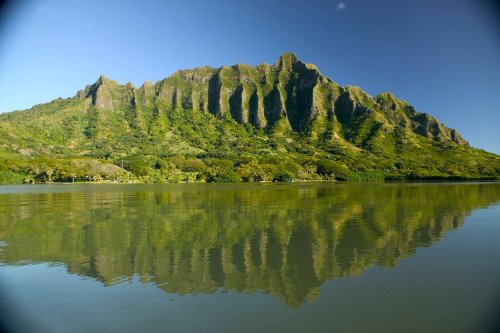 Lush green mountain reflecting in calm water under a clear blue sky.