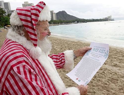 Santa Claus in a red and white striped suit reads a letter on a beach with a scenic view of the ocean and mountains.