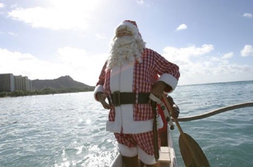 Santa Claus in a red and white outfit stands on a boat, paddling in the ocean with a sunny sky and mountains in the background.