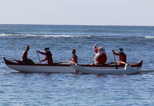 A group of people in festive attire, including Santa, paddling in a canoe on a sunny ocean.