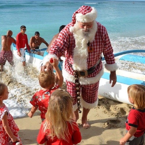 A Santa in a red and white outfit stands on a beach, surrounded by children and adults enjoying the water.