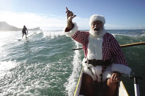 Santa Claus in a red and white outfit waves from a canoe on the ocean, with a surfer in the background.