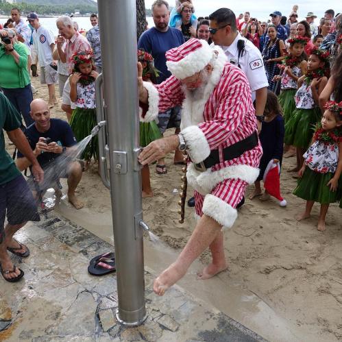 Santa Claus in a red and white suit stands by a water fountain on the beach, surrounded by festive children and onlookers.