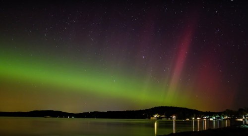 A vibrant aurora borealis lights up the night sky over a calm lake, with stars twinkling above a distant shoreline.