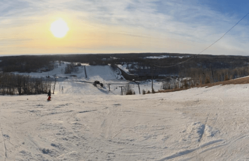 A panoramic view of a snowy ski slope at sunset, with trees and a distant road visible in the background.