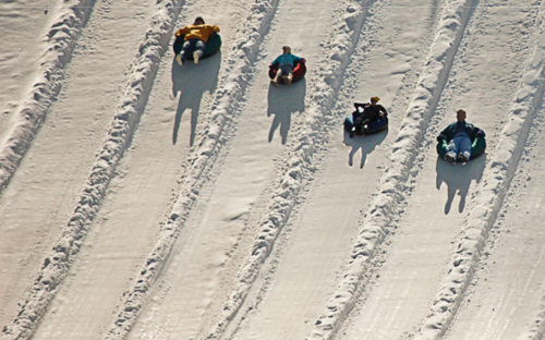 Five people sledding down a snowy hill, each on inflatable tubes, with tracks in the snow behind them.