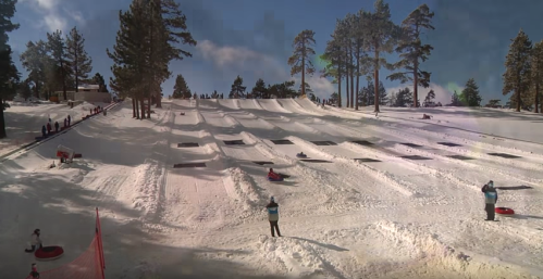 A snowy tubing hill with people sliding down and trees in the background under a clear blue sky.
