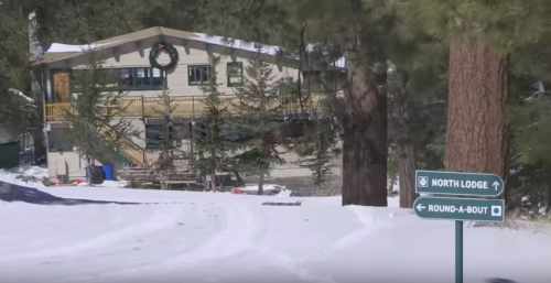 A snowy landscape featuring a lodge and directional signs for North Lodge and Round-A-Bout. Pine trees surround the area.