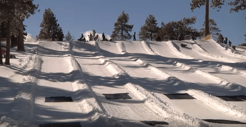 A snowy hill with multiple tubing lanes, people sliding down, and trees in the background under a clear blue sky.