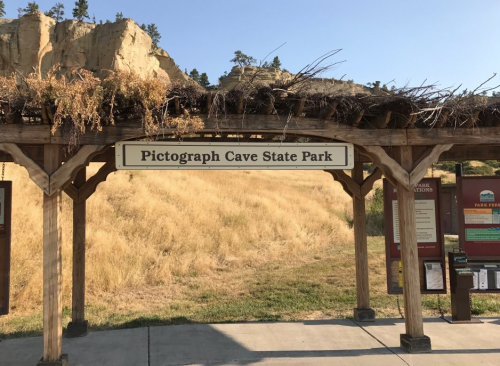 Sign for Pictograph Cave State Park, with a wooden structure and dry grass in the background.