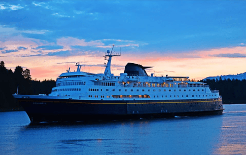 A large cruise ship sails on calm waters at sunset, surrounded by trees and mountains in the background.