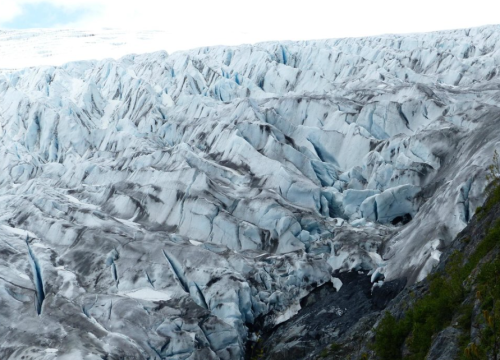 A vast glacier with blue and white ice formations, showing crevasses and rocky terrain beneath a cloudy sky.