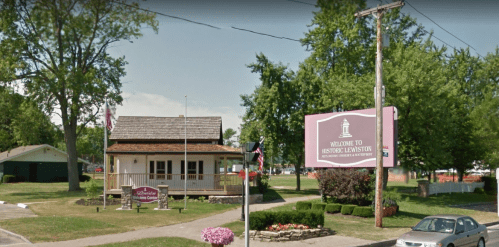 A welcome sign for Historic Lewiston in front of a small building surrounded by trees and a grassy area.