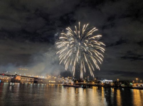 Colorful fireworks burst in the night sky over a city skyline, reflecting on the water below.
