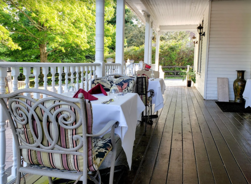 A charming outdoor dining area with white tables, elegant chairs, and lush greenery in the background.
