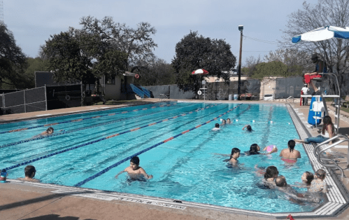 A sunny outdoor pool scene with children swimming and playing, surrounded by trees and poolside chairs.