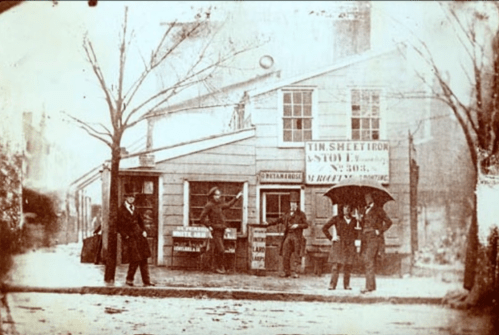Historic black-and-white photo of a storefront with people standing outside, some holding an umbrella, in a city setting.