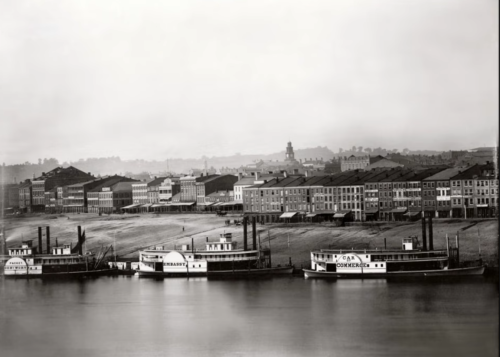 Black and white photo of a river with steamboats and a historic waterfront lined with buildings.