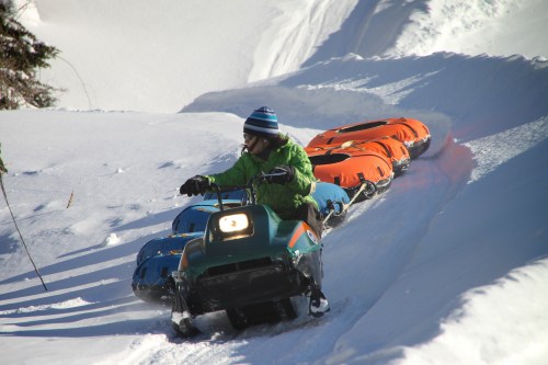 A person in a green jacket rides a snowmobile pulling colorful inflatable tubes down a snowy slope.