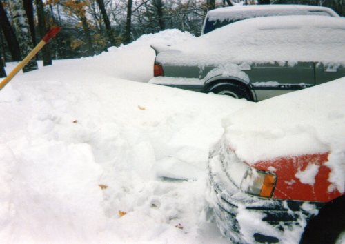 Two cars partially buried in deep snow, with a snow shovel leaning against a pile of snow in the foreground.