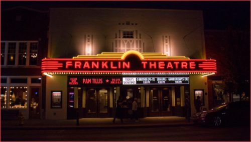 The illuminated facade of Franklin Theatre at night, featuring a marquee with event details and vibrant neon lights.