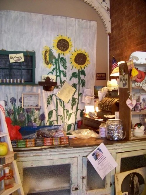 A rustic shop counter with sunflowers painted on the wall, featuring various items and a vintage cash register.