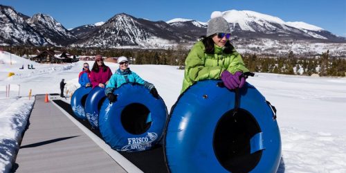 A group of people in colorful winter gear riding blue snow tubes on a snowy slope with mountains in the background.