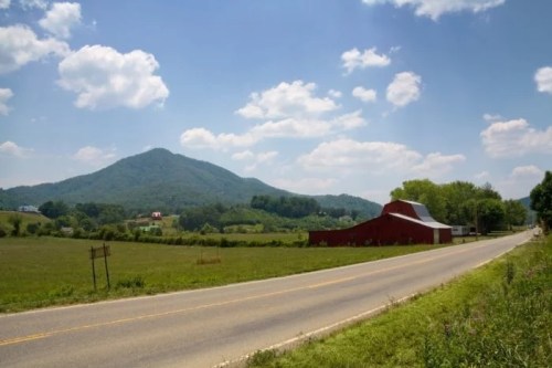 A scenic view of a rural road, red barn, and green mountains under a blue sky with fluffy clouds.