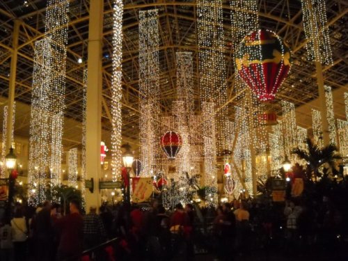 A festive indoor scene filled with twinkling lights, hot air balloons, and a crowd enjoying the holiday decorations.