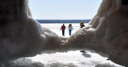 Three people walking on a frozen lake, framed by large ice formations in the foreground.