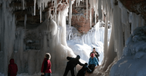People exploring a frozen cave with icicles hanging from the ceiling and snow on the ground.