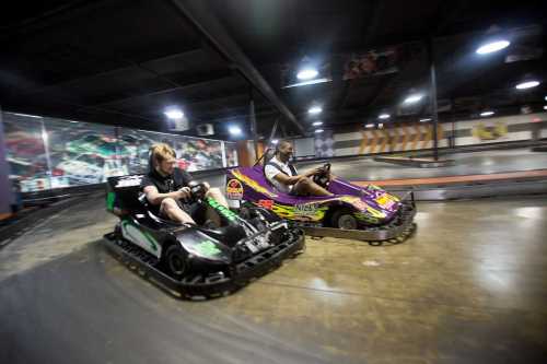 Two racers in go-karts speed around a track in an indoor racing facility.
