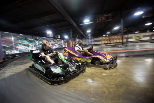 Two racers in go-karts speed around a track indoors, focused on the race ahead.